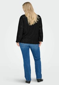 Top in pizzo nero con maniche lunghe e design con schiena scoperta, abbinato a jeans dritti blu scuro. Presenta un aspetto testurizzato e a fantasia.