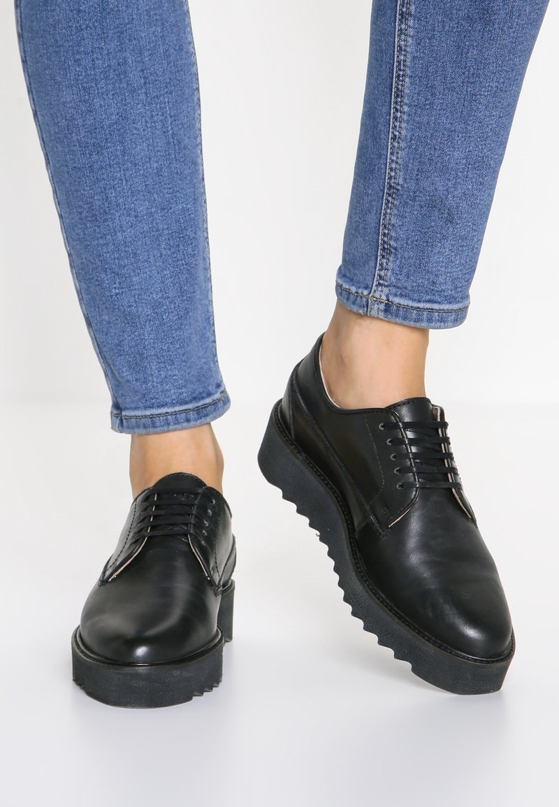Personne portant des chaussures à plateforme en cuir noir à lacets, associées à un jean skinny bleu, sur fond blanc.