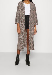 Blomsterprydd lång kimono i mörkt tyg med ljusa blommönster, vida ärmar, öppen front, bärs över en vit topp och svarta shorts, matchad med svarta stövlar.