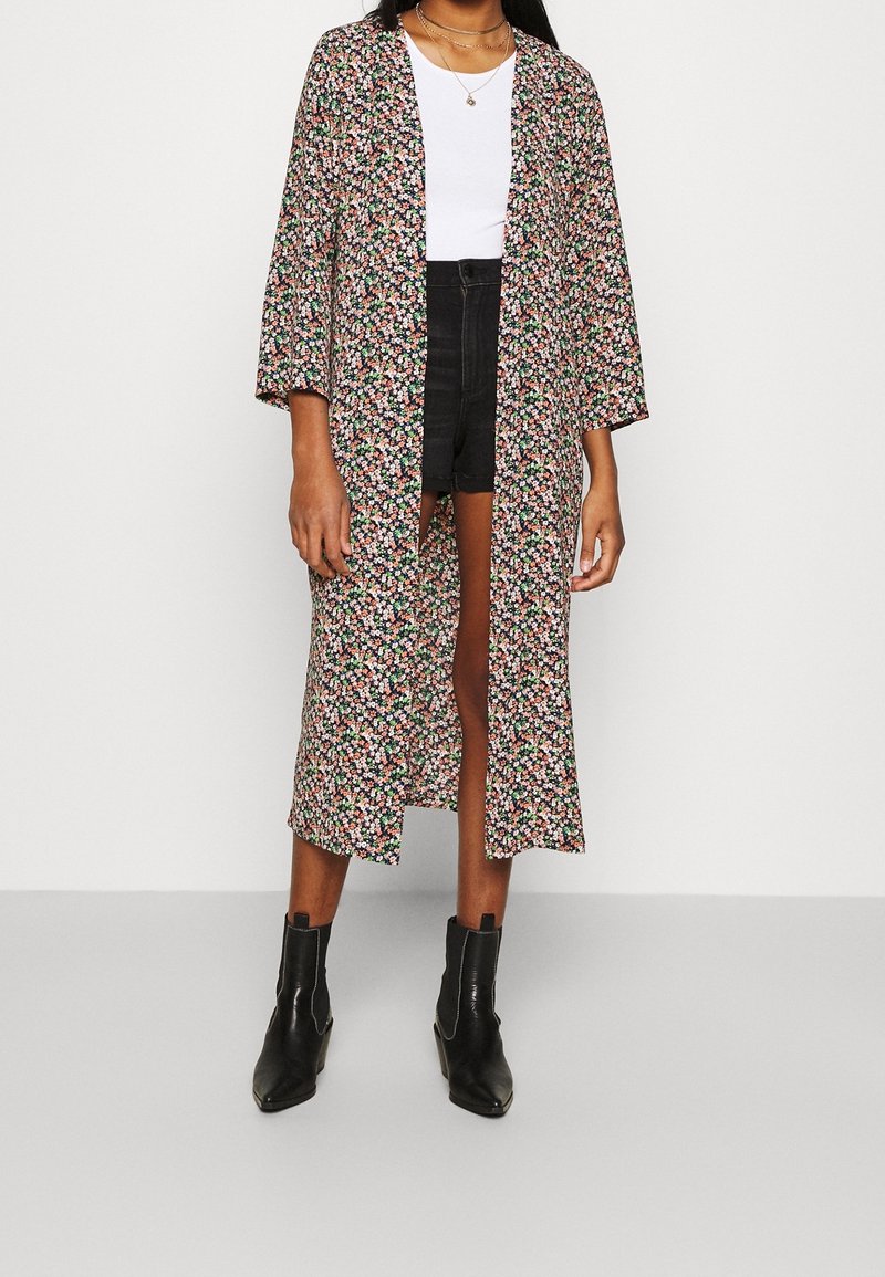 Blomsterprydd lång kimono i mörkt tyg med ljusa blommönster, vida ärmar, öppen front, bärs över en vit topp och svarta shorts, matchad med svarta stövlar.