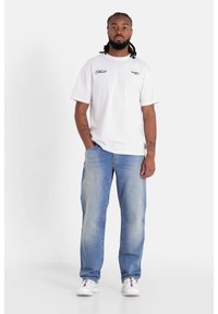 T-shirt bianca in cotone con loghi stampati, abbinata a jeans in denim azzurro chiaro. Il modello indossa sneakers bianche; presenta una vestibilità rilassata e un leggero effetto invecchiato.