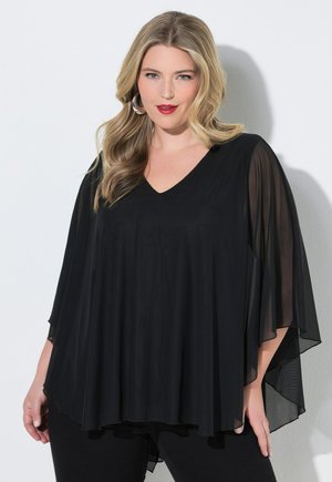 Lyshåret kvinde med løse bølger iført creoløreringe og en sort flydende top med gennemsigtige ærmer, stående foran en hvid baggrund.