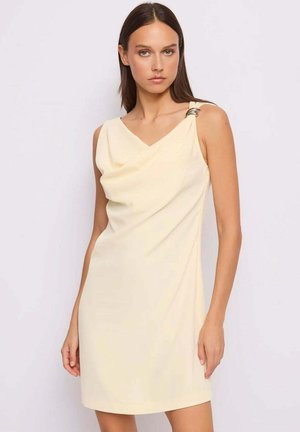 Donna che indossa un abito crema senza maniche con scollo drappeggiato e dettaglio argentato sulla spalla sinistra, in piedi davanti a uno sfondo neutro.