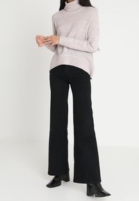 Hellrosa gestrickter Pullover mit hohem Kragen, kombiniert mit hoch taillierter schwarzer Schlaghose und schwarzen Stiefeletten mit Absatz.