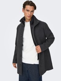 Grauer Wollmantel mit hohem Kragen, Fünf-Knopf-Verschluss und schwarzem Innenfutter. Das Modell trägt ihn über einem weißen, strukturierten Pullover.