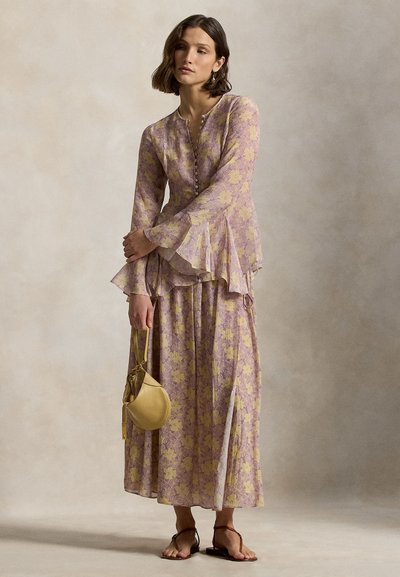 Frau trägt ein langärmliges Kleid mit floralem Muster in Lavendel und Gelb, hält eine kleine gelbe Handtasche und trägt Sandalen mit Knöchelriemen.