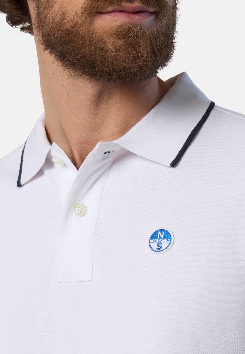Polo blanco con tejido texturizado, que presenta un cuello azul marino, tapeta de tres botones y un logo redondo azul en el lado izquierdo del pecho.