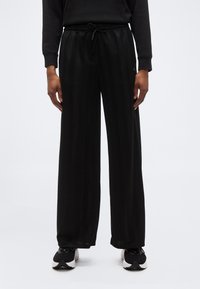 Άτομο που φοράει μαύρο παντελόνι με φαρδιά πόδια και διακριτικές κάθετες ρίγες, μαύρο μακρυμάνικο τοπ και μαύρα και άσπρα αθλητικά παπούτσια σε λευκό φόντο.