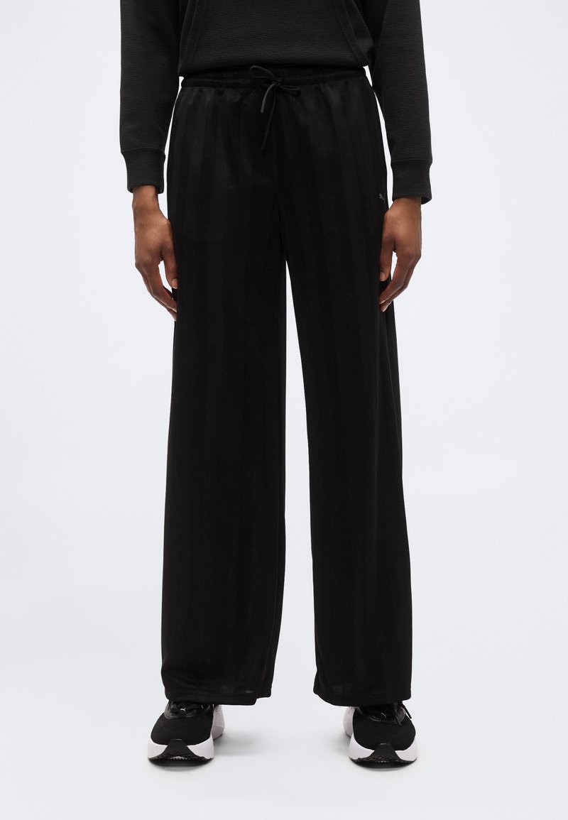 Άτομο που φοράει μαύρο παντελόνι με φαρδιά πόδια και διακριτικές κάθετες ρίγες, μαύρο μακρυμάνικο τοπ και μαύρα και άσπρα αθλητικά παπούτσια σε λευκό φόντο.