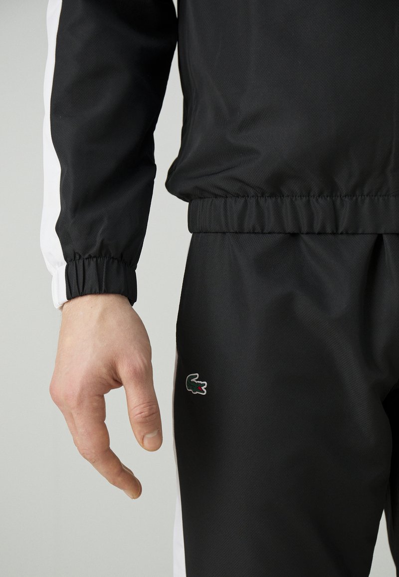 Ingrandimento della mano di una persona e di un completo sportivo nero con strisce bianche e un piccolo logo di coccodrillo verde sui pantaloni.