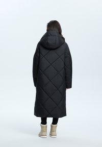 Langer schwarzer Daunenmantel mit Kapuze, ausgestattet mit einem rautenförmigen Steppmuster und einer lockeren Passform, getragen über schwarzen Leggings und beigen Stiefeln.