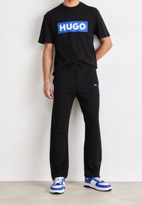 Schwarzes T-Shirt mit blauer "HUGO"-Logo, kombiniert mit schwarzen Jogginghosen mit Kordelzug und blau-weißen Sneakers, vor einem hellen Hintergrund stehend.