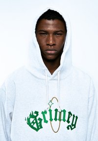 Sudadera gris con capucha ajustable, que presenta texto y acentos bordados en verde. La tela parece suave con una textura lisa.