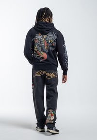 Felpa nera con una grafica di un drago colorato sul retro, abbinata a jeans scuri con design ricamati e il marchio "Ed Hardy".