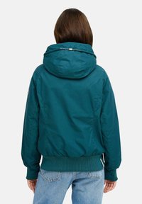 Giacca leggera di colore verde acqua con cappuccio, polsini e orlo a coste, e dettagli di cucitura sottili. La vista posterior mostra un design elegante e affusolato.