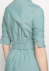 Abito a strisce verdi e bianche con maniche corte. Caratterizzato da una texture arricciata sulle spalle e una vita a cintura per una silhouette definita.