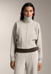 Svetlo šedá bundá na zips s vysokým golierom a rebrovanými detailmi, s pevnou siluetou a tmavohnedým pásom. Mäkká textúra po celom povrchu.