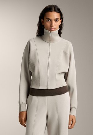 Hellgraue Zip-Up-Jacke mit hohem Kragen und gerippten Akzenten, mit einer strukturierten Passform und einem dunkelbraunen Bund. Weicher Griff über die gesamte Fläche.