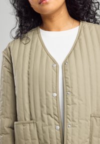 Chaqueta acolchada beige con diseño de escote en V, botones de presión de metal y dos bolsillos laterales. La tela tiene una textura suave con costuras visibles.