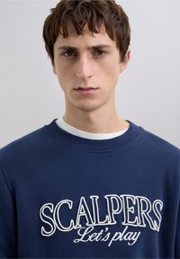 Sudadera azul marino de algodón, con texto bordado en blanco "SCALPERS Let's play", con cuello acanalado y textura suave.