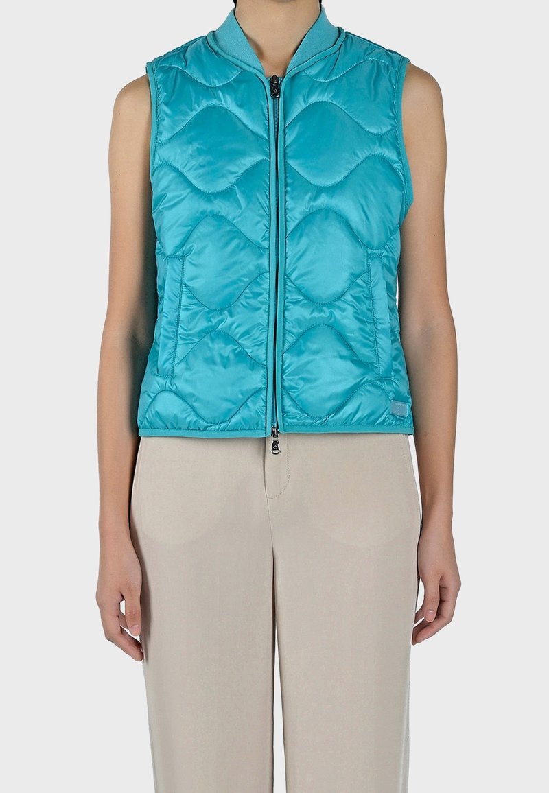 Gilet sans manches matelassé en turquoise vif avec un motif piqué, col montant et fermeture éclair sur le devant, associé à un pantalon beige.