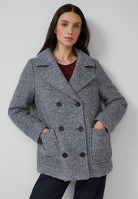 Femme aux longs cheveux noirs portant un manteau texturé gris à double boutonnage avec de grands revers et des boutons noirs, les mains dans les poches avant, fond neutre.