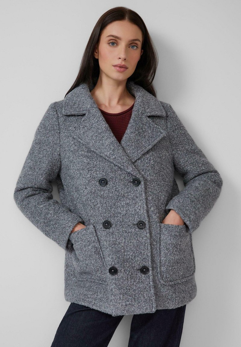 Femme aux longs cheveux noirs portant un manteau texturé gris à double boutonnage avec de grands revers et des boutons noirs, les mains dans les poches avant, fond neutre.