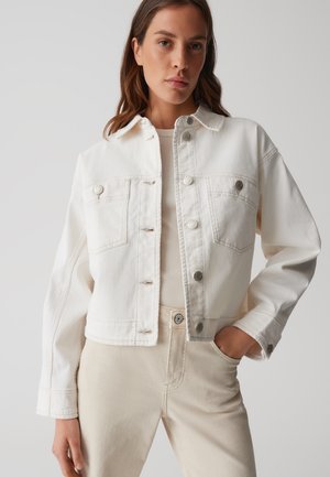 Witte denimjack met knoopsluiting aan de voorkant, twee borstzakken en lange mouwen; gedragen over een lichtbeige top en lichtbeige broek.