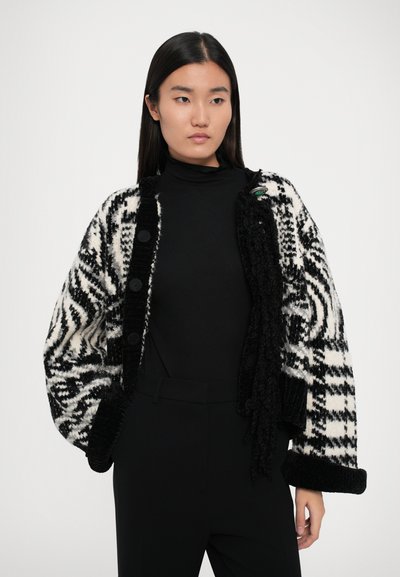 Cardigan noir et blanc à motifs avec une finition texturée, design oversize, et trois grands boutons, porté sur un col roulé noir.