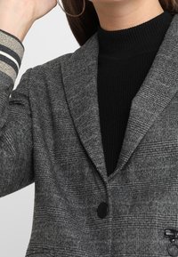 Szary marynarka w kratę z klapami z wycięciem i pojedynczym guzikiem. Pod spodem czarny prążkowany golf o fakturowanej tkaninie.