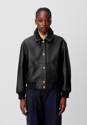 Persona rivolta in avanti indossa una giacca di pelle nera con bottoni dorati, camicia beige e pantaloni blu navy su uno sfondo chiaro e semplice.