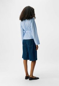 Camicetta a righe blu chiaro con maniche lunghe, abbinata a bermuda di jeans scuri. Le scarpe sono ballerine nere. Vista posteriore del modello.