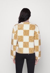 Fuzzy, rutigt jacka i crème och beige, med en mjuk textur, långa ärmar och en kort klippning. Bakvy med rund halsringning.