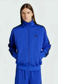 Jauns cilvēks ar gariem taisniem matiem valkā zilu Adidas sporta jaku un atbilstošas šortus, stāvot pret vienkāršu gaišu fona.