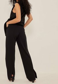 Pantaloni neri a gamba larga con fessure laterali, caratterizzati da una texture liscia e una vestibilità sartoriale. Indossati con un top senza maniche nero, dal design minimal.