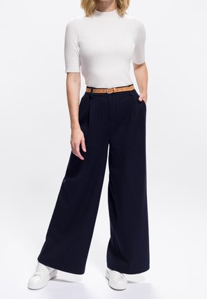 Femme portant un haut blanc à manches courtes, un pantalon large à rayures bleu marine avec une ceinture brune et des baskets blanches, se tenant devant un fond blanc uni.