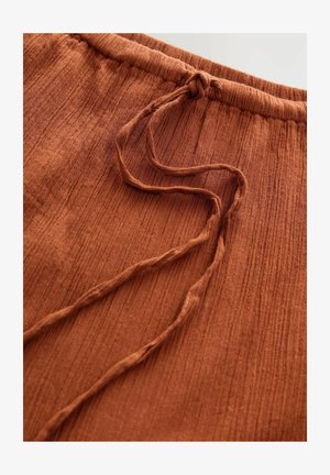 Tessuto di colore ruggine con texture a righe verticali, caratterizzato da una vita elastica e un cordoncino per una vestibilità regolabile. Materiale morbido.
