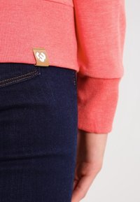 Felpa corallo con texture a coste, maniche aderenti e un piccolo etichetta a marchio intrecciata. Abbinata a jeans blu scuro, leggermente elasticizzati.