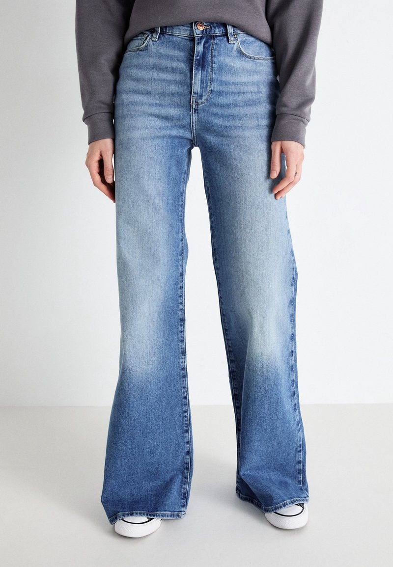 Jean évasé en denim bleu clair à taille haute, avec une texture délavée et un design classique à cinq poches, porté avec des baskets blanches.