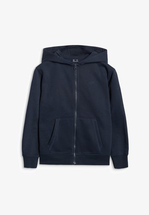 Fille aux longs cheveux bruns porte un t-shirt court blanc, un sweat à capuche zippé bleu marine, un pantalon de jogging assorti et des baskets noires et blanches, debout en souriant.
