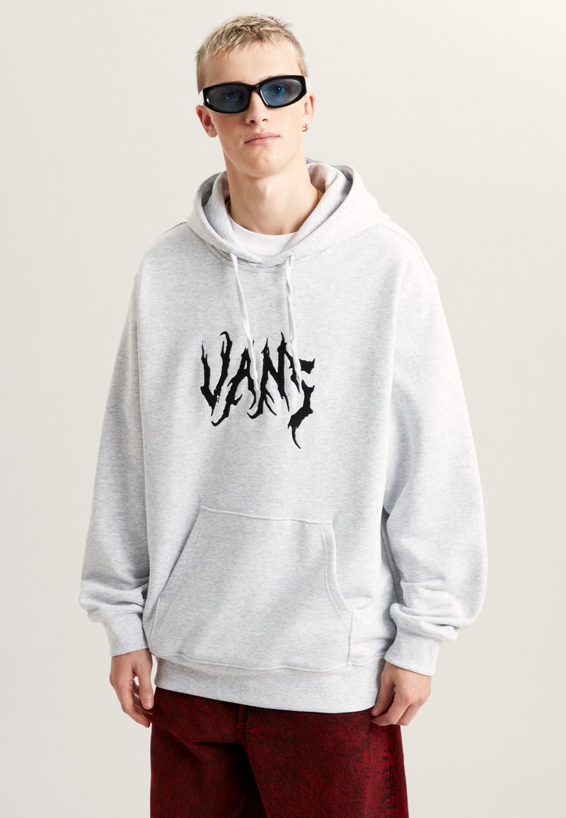 Haut à capuche gris avec poche frontale, logo "VANS" noir et capuche à cordon réglable ; associé à un pantalon rouge à motifs et des lunettes de soleil noires.