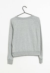 Šedá bavlněná mikina s kruhovým výstřihem a žebrovanými manžetami. Má raglánové rukávy a hladký, texturovaný povrch. Visí na černém háčku.