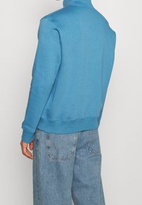 Sudadera azul con cuello alto, puños y dobladillo acanalados. Hecha de una tela suave, se muestra combinada con unos vaqueros anchos de color azul claro.
