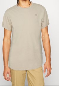 Svetle béžové tričko s krátkým rukávem vyrobené z bavlny, s kulatým výstřihem a malým logem na levém prsu. Hladká textura.