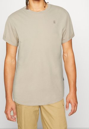 T-shirt beige chiaro a maniche corte in cotone, con scollo rotondo e un piccolo logo sul petto sinistro. Texture liscia.