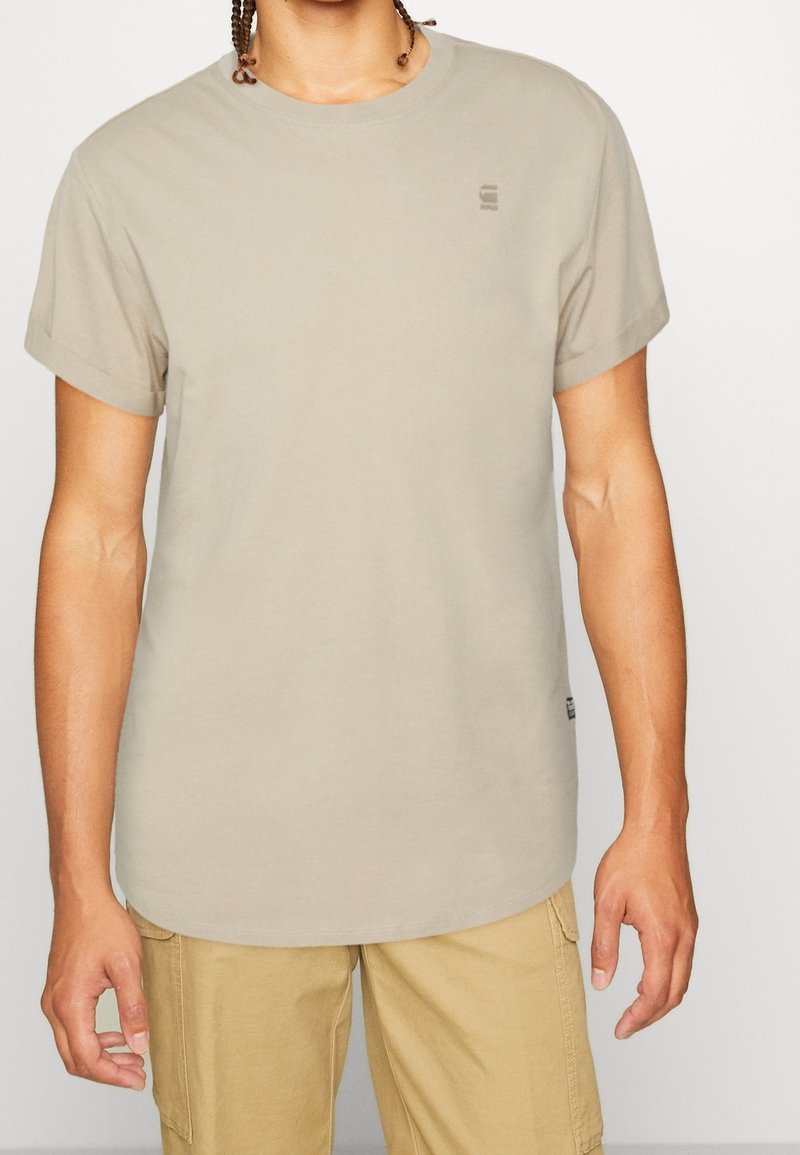 Svetle béžové tričko s krátkým rukávem vyrobené z bavlny, s kulatým výstřihem a malým logem na levém prsu. Hladká textura.