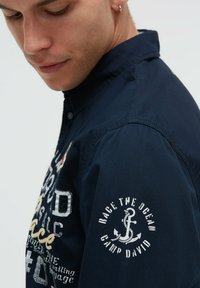 Marineblaue Hemdjacke mit bestickten Grafiken, darunter ein Anker und Text auf dem Ärmel. Der Stoff wirkt strukturiert und strapazierfähig.