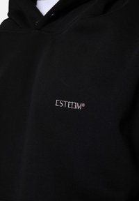 Haut à capuche noir en tissu doux, avec un petit logo brodé blanc "ESTEEM" sur le devant. Design simple avec une coupe classique.