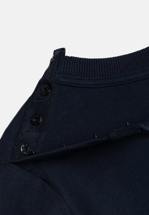 Dunkelblaues geripptes Halsbündchen mit drei schwarzen Druckknöpfen, die teilweise an der Schulter eines Strickoberteils geöffnet sind.