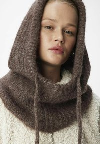 Un col en tricot marron fabriqué en fil texturé et flou avec un cordon de serrage. Porté par-dessus une veste crème duveteuse avec un col arrondi.